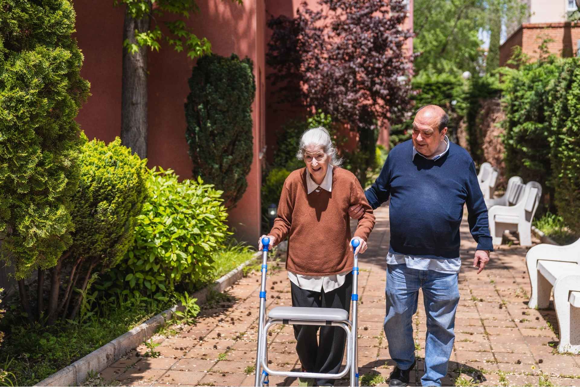 A senior woman using her walker outdoors, with a senior man by her side providing support.